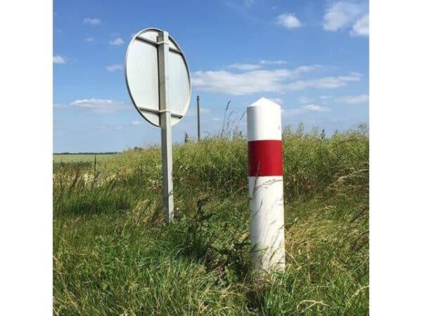 Photo d'une balise de virage et intersection J1-J3 blanc avec un bandeau rouge, dans l'herbe, derrière un panneau