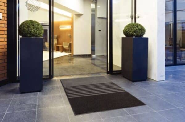 Photo d'un tapis d'accueil 3 en 1 (grattant, grattant et absorbant, absorbant et anti-poussière) noir et gris avant un SAS d'entrée d'immeuble
